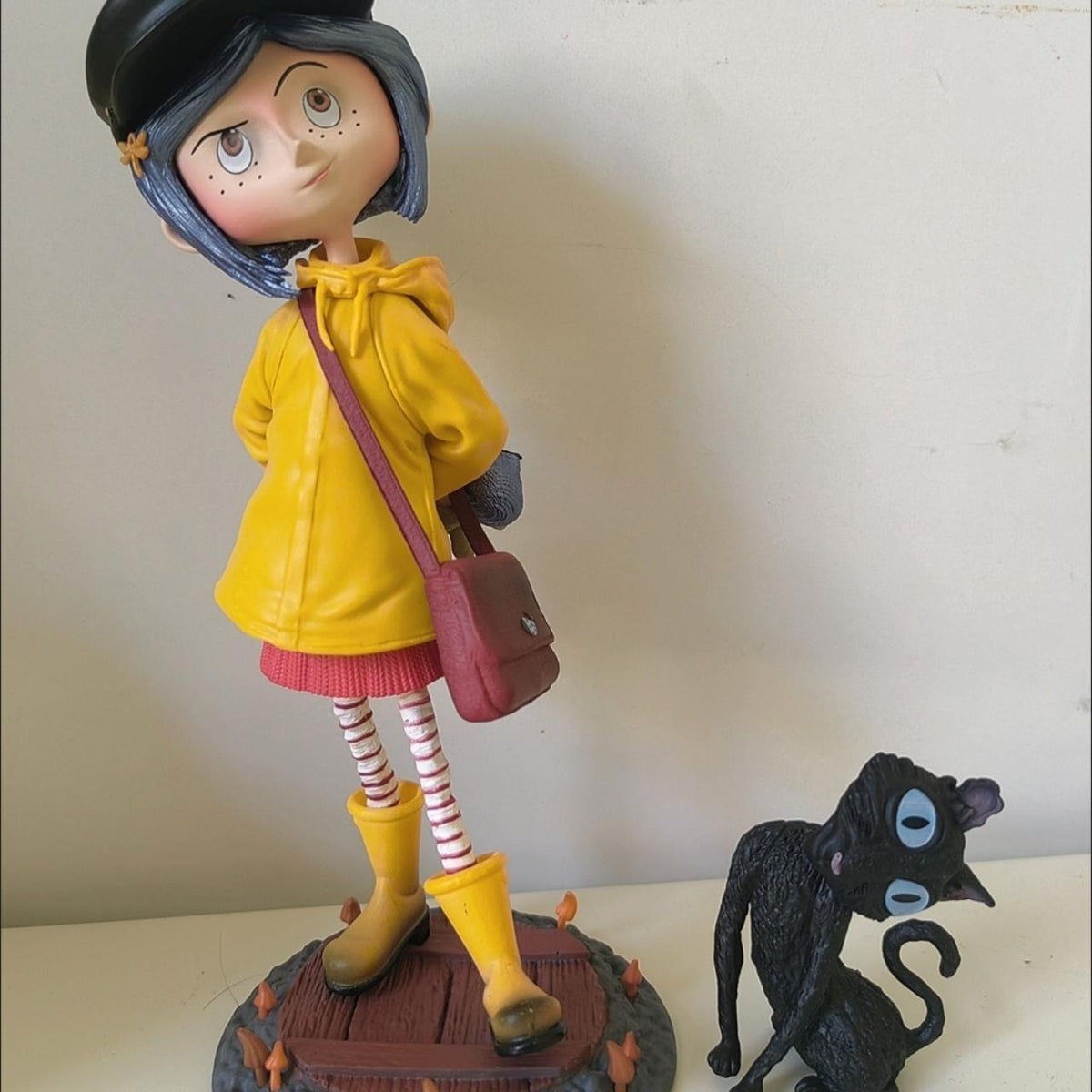 Coraline raincoat girl figure standing on its sculpted base — full-length view with the black cat companion to the right
