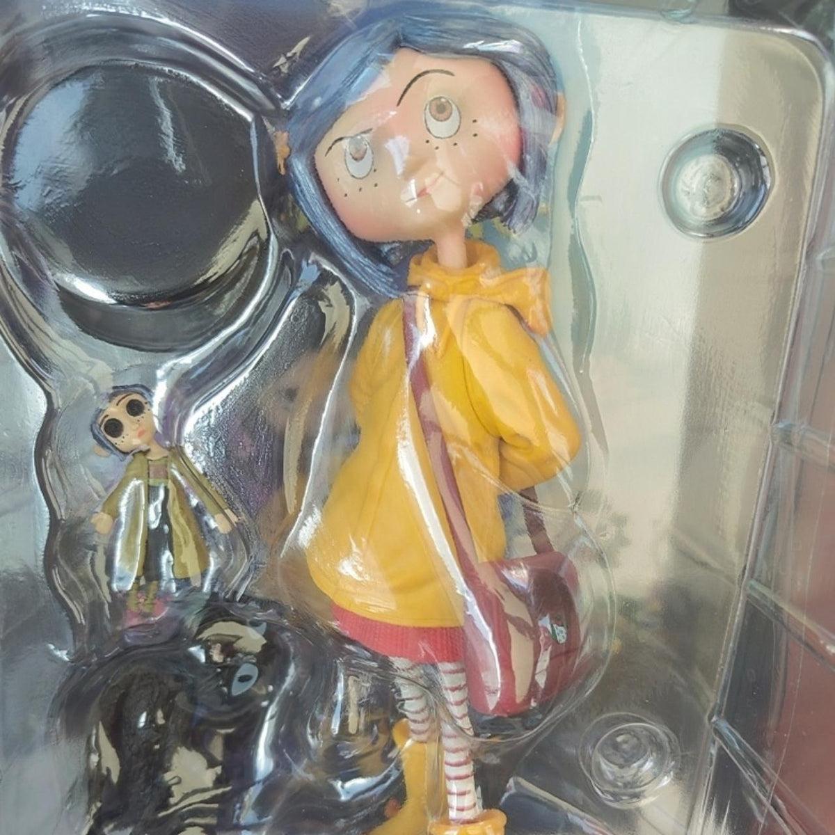 Coraline raincoat girl figure still sealed in its original display box and blister tray — yellow raincoat, blue bob and striped tights visible through the plastic