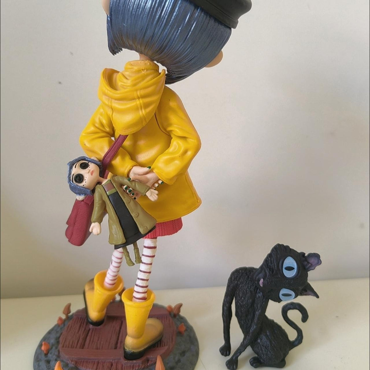 Side view of the Coraline raincoat girl figure on its grass base with the black cat companion sitting beside it
