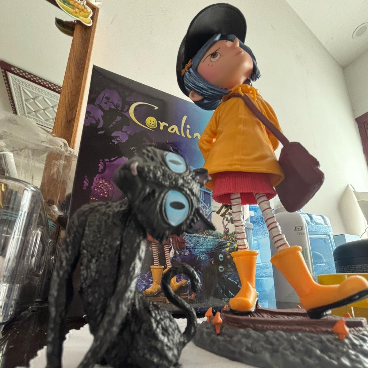 Coraline raincoat girl figure displayed on its bridge base beside the black long-legged cat, with a Coraline backdrop card behind