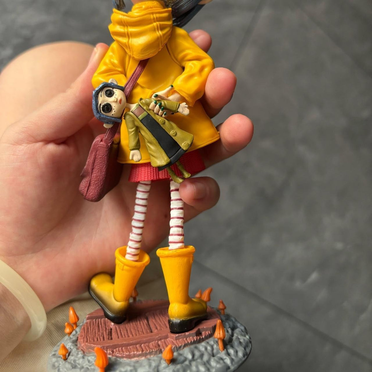 Back view of the Coraline raincoat girl figure showing the hooded yellow raincoat, red skirt and the miniature shoulder figure