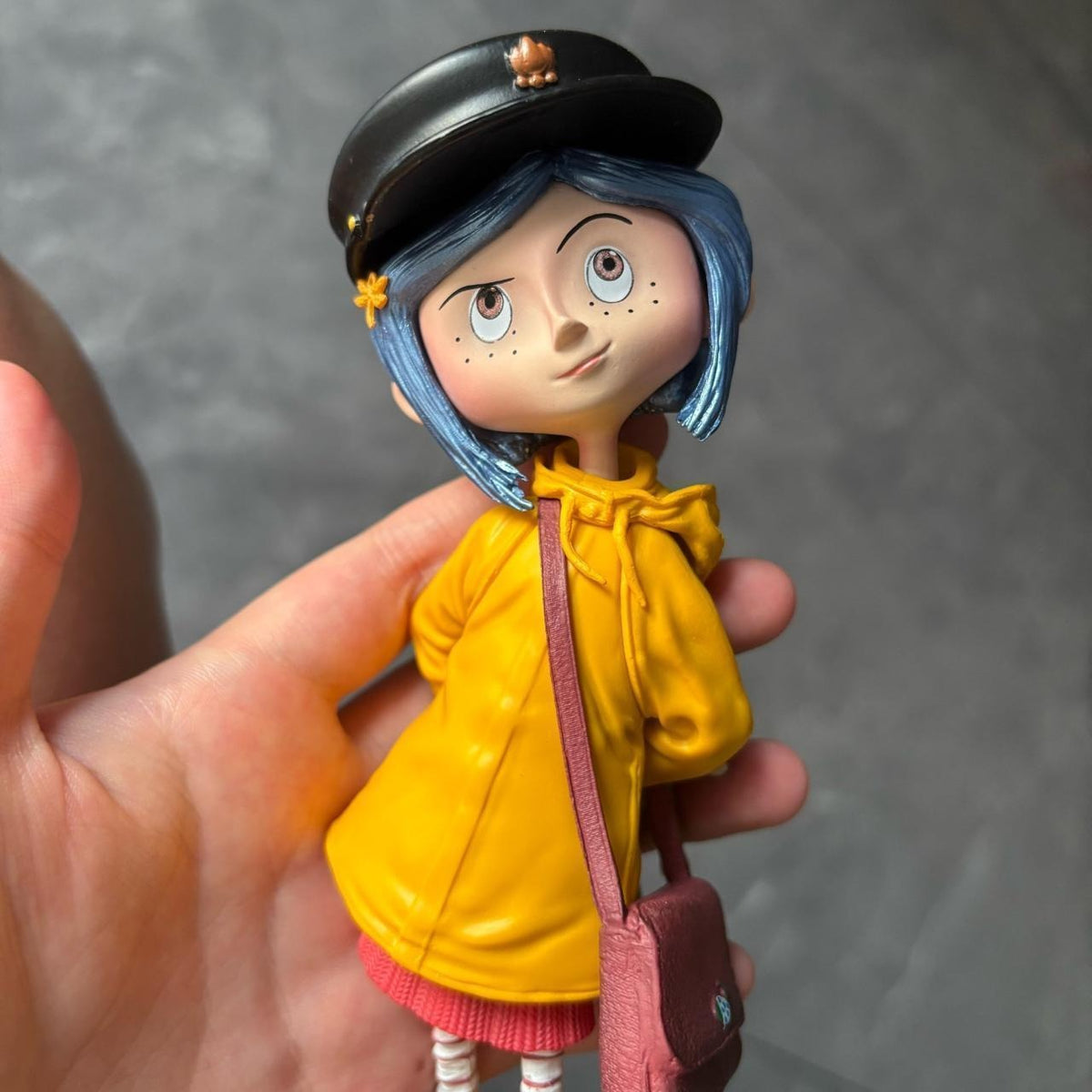 Coraline raincoat girl figure held in hand for scale — black cap with flower pin, blue bob and yellow bow-collar raincoat